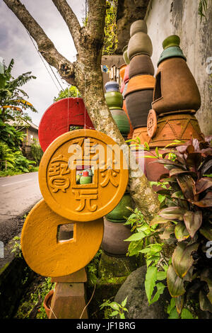 Three Iching lucky coins hang from a tree in front of rows of colourful pottery items, stacked on top of each other. Stock Photo