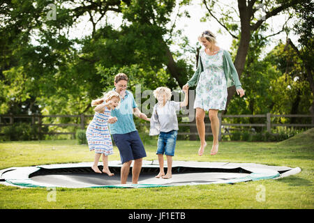 Man, woman, boy and girl holding hands, jumping on a trampoline set into the ground in a garden. Stock Photo