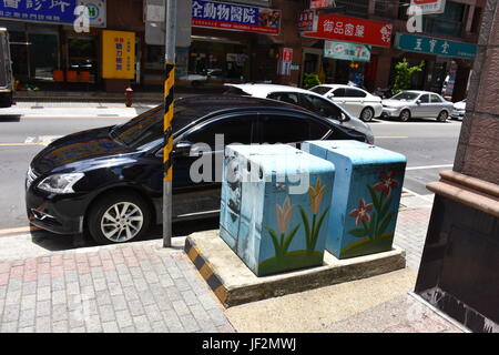 A black car parked near city power boxes that were painted blue with tall flowers on them to make them more pleasing to the eye. Stock Photo