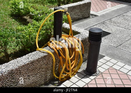 Long yellow garden hose rolled up around a faucet near bushes and grass on stone wall. Stock Photo