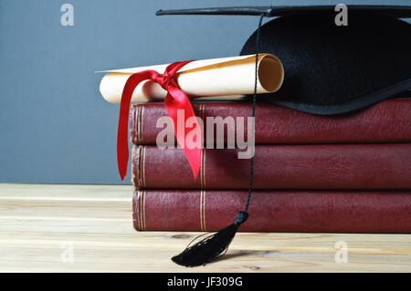 Graduation mortarboard and scroll tied with red ribbon on top of a stack of old, worn books on a light wood table.  Grey background. Stock Photo