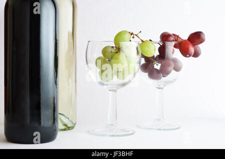 Two bottles of wine on left side of two wine glasses containing red and white grapes. Stock Photo