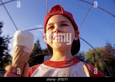 Young girl (7 years old), Little League Baseball player, eating ice cream after game, sunset light, portrait USA Stock Photo