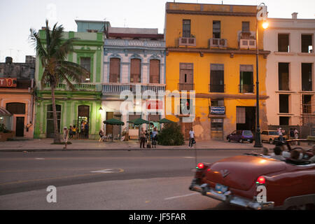 At dusk, people stand on the street as a classic American car drives by in downtown Havana. Stock Photo