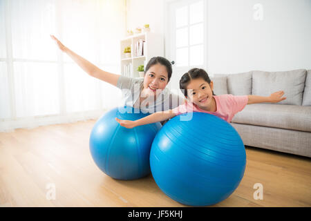 young mixed race asian chinese mother and her daughter lying on the big softness ball and opening hands as flying cheerful doing yoga exercise at home Stock Photo