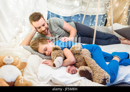 Smiling father looking at cute little son sleeping with teddy bear in blanket fort Stock Photo