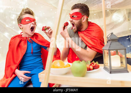 Happy father and son in superhero costumes eating strawberries in blanket fort Stock Photo