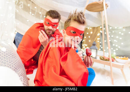 Happy father and son in superhero costumes eating strawberries in blanket fort Stock Photo