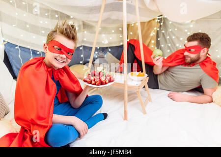 Happy father and son in superhero costumes eating fruits in blanket fort Stock Photo