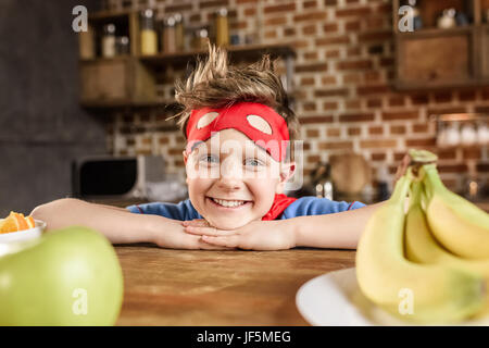 smiling boy in red superhero costume sitting in kitchen and looking at camera Stock Photo