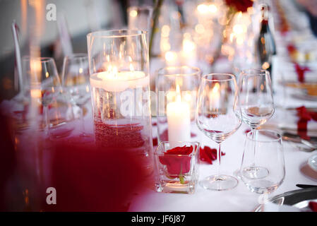 Romantic candlelit dinner table setting with elegant glassware and red flowers over a white tablecloth in a close up view Stock Photo