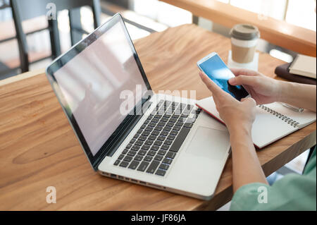 Asian hipster male using smartphone for social network while working with laptop computer in coffee shop. Freelancer lifestyle and online activity Stock Photo