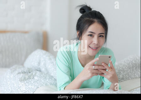 Young attractive Asian woman wear green casual cloths using smartphone for social network while laying down on bed. Lazy girl activity on sunday morni Stock Photo