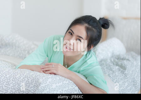 Young attractive Asian woman wear green casual cloth smiling while laying down on bed. Lazy girl activity on sunday morning. Stock Photo
