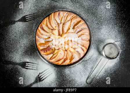 Apple pie in a portioned cast-iron frying pan, on a black stone table sprinkled with powdered sugar, with traces of forks. The concept of food and gue Stock Photo