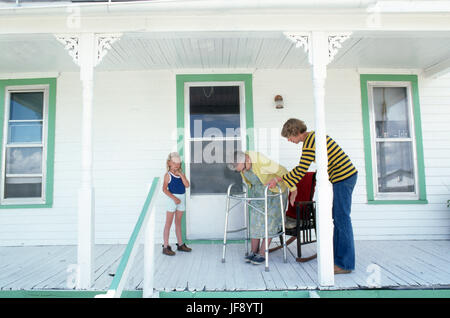 3 generations of women daughter helping elderly mother to sit in her rocker on her front porch while young daughter looks on Stock Photo