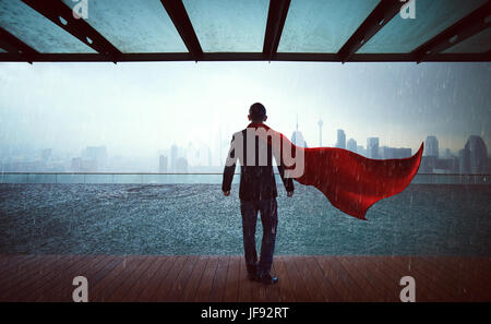 Businessman in a suit and cape hero stand at sky garden ,victory concept. cityscape background , raining day. Stock Photo