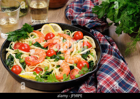 Pasta with fried prawns, peas, tomatoes and spinach in a frying pan, on a table with cider glasses. Stock Photo