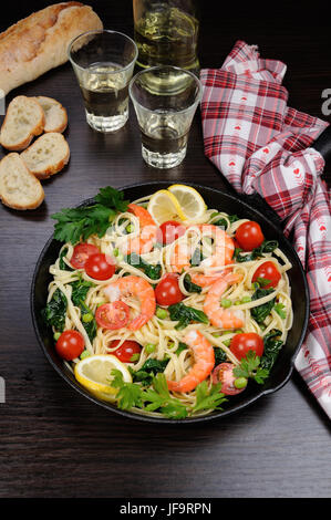 Pasta with fried prawns, peas, tomatoes and spinach in a frying pan, on a table with cider glasses. Stock Photo