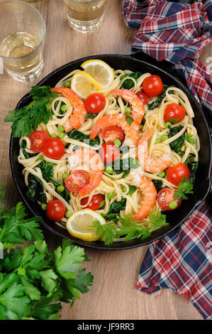 Pasta with fried prawns, peas, tomatoes and spinach in a frying pan, on a table with cider glasses. Stock Photo