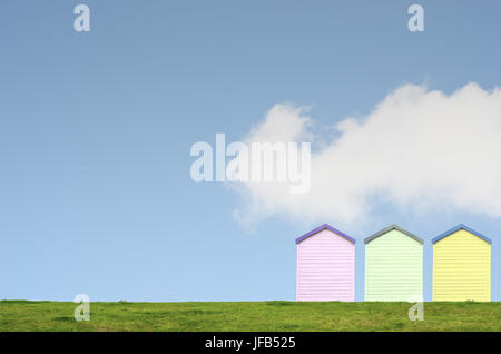 A row of three colourful beach huts on top of a grassy hill against a bright blue sky with white fluffy cloud. Copy space above and left. Stock Photo