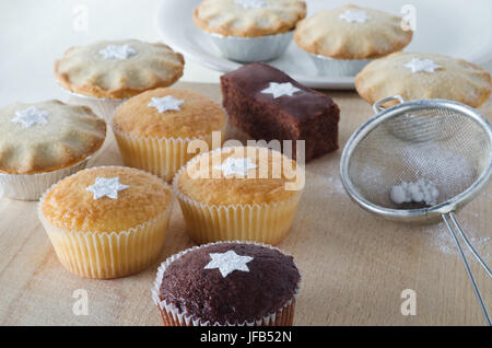 A group of Christmas cakes and mince pies, decorated with star shapes from sifted icing sugar, on a wooden chopping board with plate in background, an Stock Photo