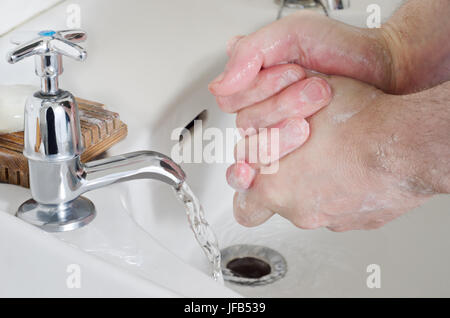 Male hands being washed in old china sink with water running from stainless steel tap, and a bar of soap resting on a wooden soap dish. Stock Photo