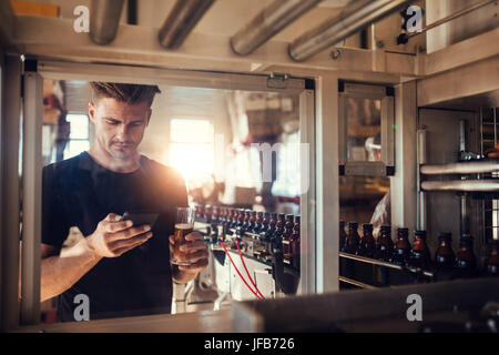 Young man with a glass of beer at brewery plant using mobile phone. Beer factory owner at bottling machine reading text message on his smart phone. Stock Photo