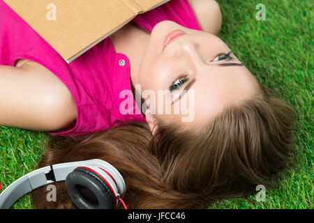 Young beautiful dreamy woman laying on grass in the park with a book and headphones beside her. Stock Photo