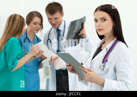 Portrait of brunette female medicine doctor holding document folder with three her colleagues looking at cardiogram and x-ray picture at background. H Stock Photo