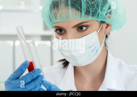 Medical doctor in protective gloves and surgical mask and hat comparing two flasks with dark red liquid in laboratory. Scientific research, healthcare Stock Photo