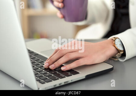 Close up view of female student at workplace holding cup of tea and working at laptop. Woman writing letter, list, plan, making notes, doing homework. Stock Photo