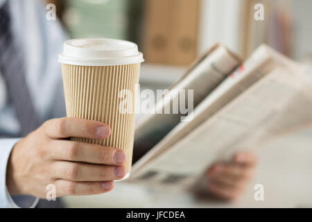 Closeup view of businessman hand holding cardboard coffee cup and reading morning newspaper. Financial or sports news, business analytics, coffeebreak Stock Photo