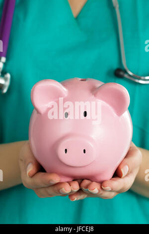 Female medicine doctor wearing green uniform holding funny pink piggy bank. Doctor hands close up. Medical service economy, health care savings and in Stock Photo