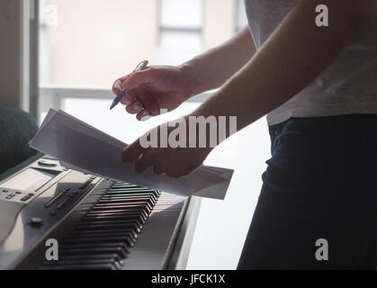 Musician brainstorming and innovating new song ideas at piano by the window. Composer writing notes to paper or planning arrangement. Stock Photo