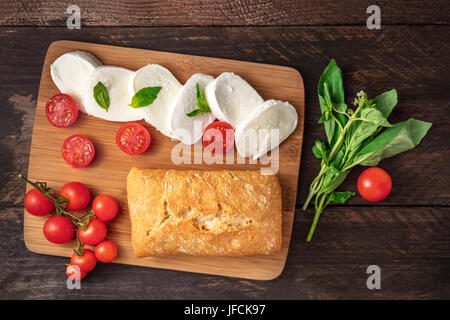 An overhead photo of buffalo mozzarella cheese with cherry tomatoes, green basil leaves, the ingredients of the Caprese salad, and a ciabatta, on a da Stock Photo