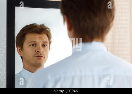 Portrait of unsure young businessman with unhappy face looking at the mirror. Man preparing for important meeting, new job interview or dating. Diffic Stock Photo