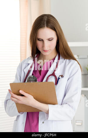 Portrait of young female doctor holding clipping pad and reading patient history or prescribing some medicines. Healthcare and medical concept. Stock Photo