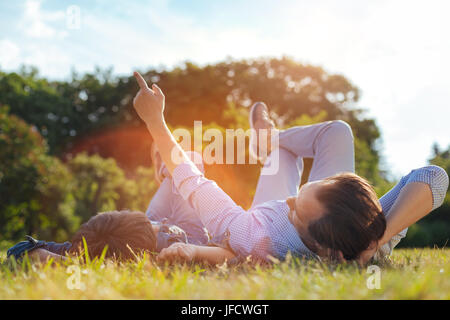 Look at that. Intelligent creative nice man pointing at the sky and explaining something while lying on the grass in a park and talking Stock Photo
