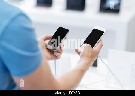 Which one is better. Scaled up shot of a man standing at a store display and holding two different models of smartphone while making a decision and ch Stock Photo