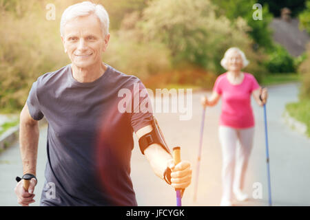 Healthy way of life. Delighted joyful nice man holding walking poles and practicing sport while being in the park Stock Photo
