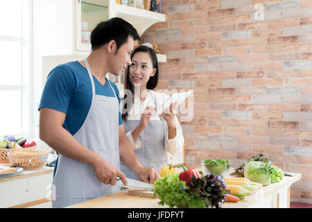 Asian beautiful young husband and wife searching menu recipe in digital tablet in kitchen at home. Happy love couple and technology concept. Stock Photo