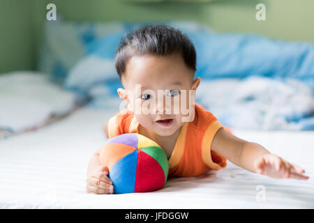 Asian adorable baby boy playing with colorful rainbow ball toy in white sunny bedroom at home. Stock Photo