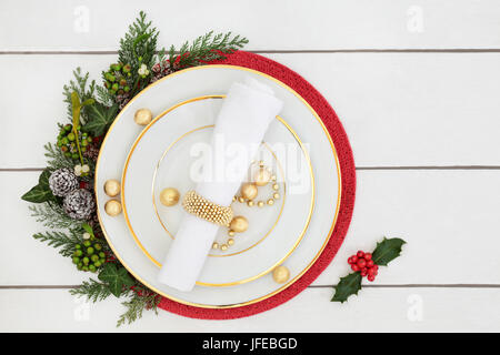 Christmas dinner table setting with porcelain plates, napkin, decorations and foil wrapped chocolates, with holly, mistletoe, ivy and cedar leaves. Stock Photo