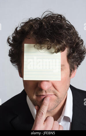 A curly haired man in a suit with shirt slightly open and no tie, resting finger on chin in a contemplative manner, with a blank, lined yellow sticky  Stock Photo