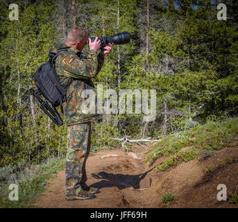 Wildlife, nature man photographer in camouflage outfit with a backpack and tripod standing on a mountain forest trail and shooting, taking pictures Stock Photo