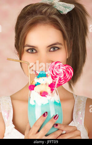 Close up of pretty cute woman holding a tasty blue milk shake with a candy heart on top, trendy clothes, studio fashion in a pink background. Stock Photo
