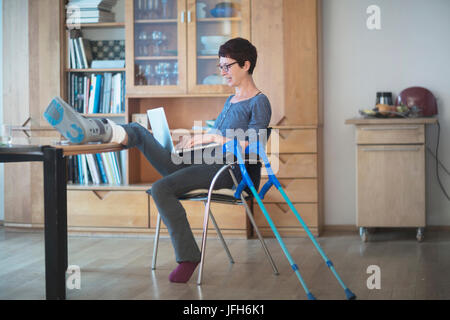 Woman resting her broken leg on table and using laptop on chair Stock Photo