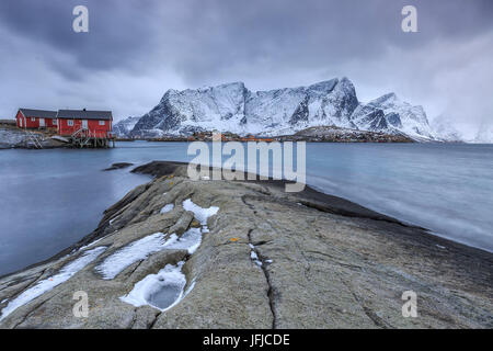 Typical red houses in the Hamnøy landscape with its cold sea and snow capped peaks, Lofoten Islands Norway Europe Stock Photo