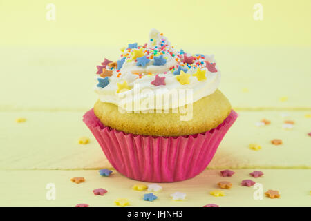 Single vanilla sponge birthday cupcake frosted and coated with star candy sprinkles on pale yellow background Stock Photo